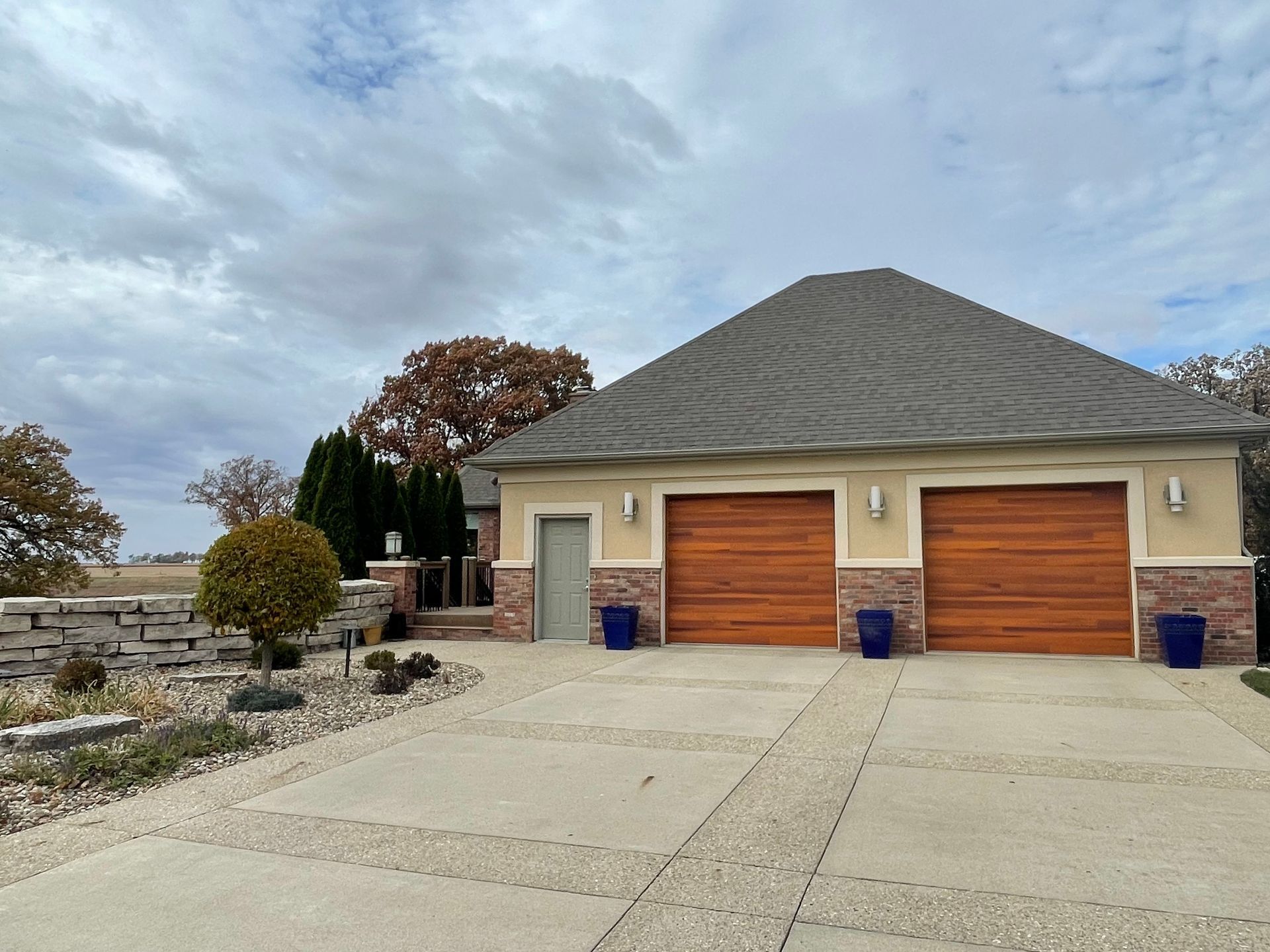 A two-car garage with wood-grain doors, beige stucco walls, and stone accents, set in a landscape with a paved driveway.