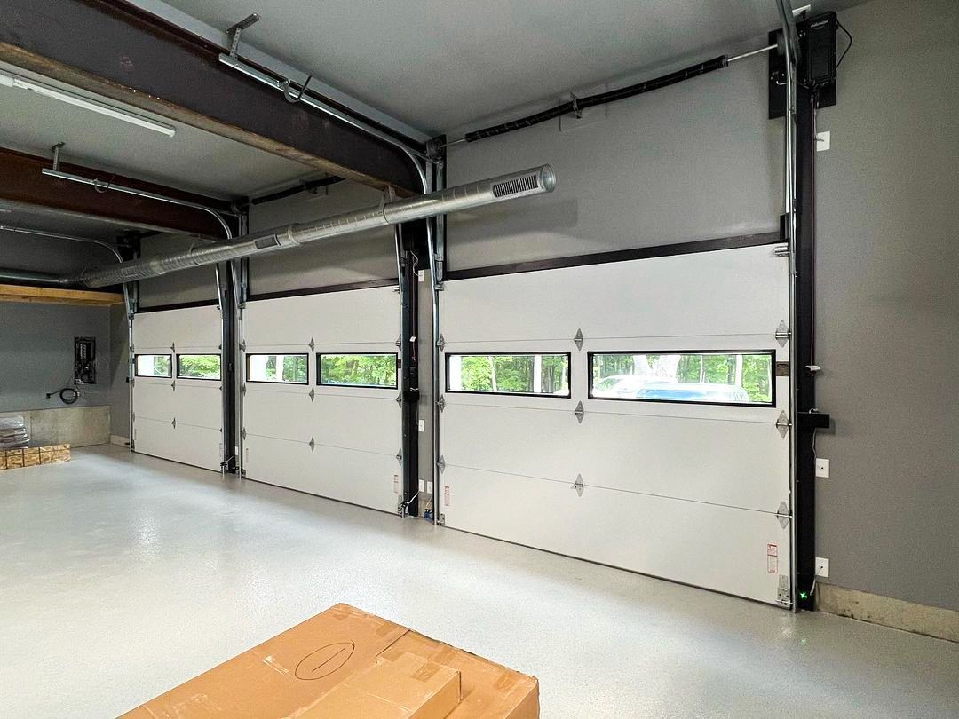 Three industrial garage doors with horizontal windows installed side-by-side in a brightly lit, spacious workshop.