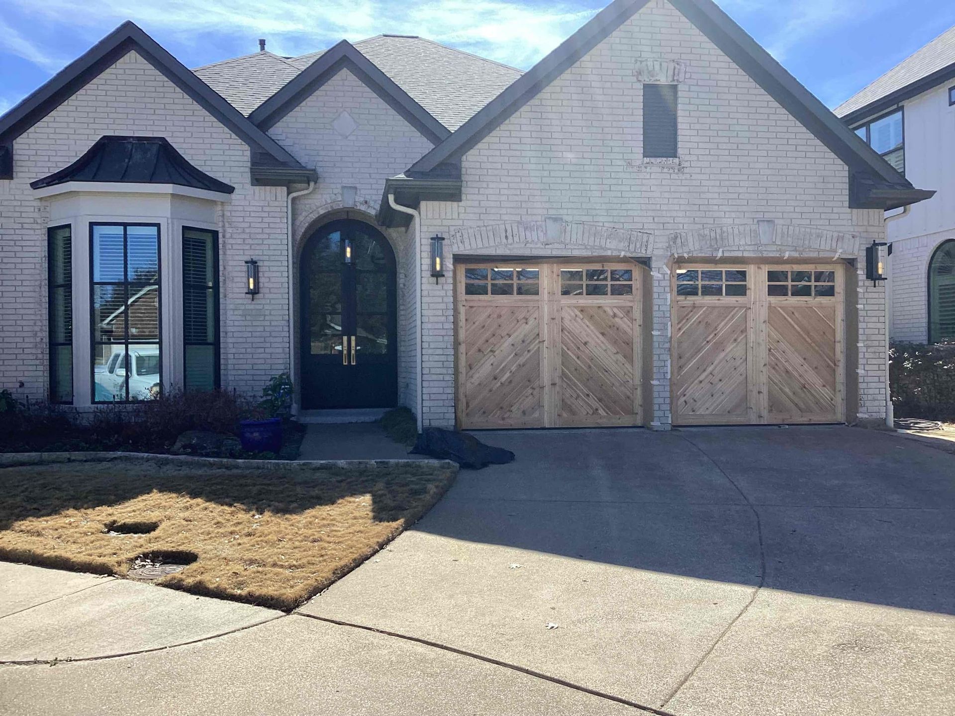 A single-story, light-colored brick house with an arched entryway and two wooden garage doors with diagonal paneling.