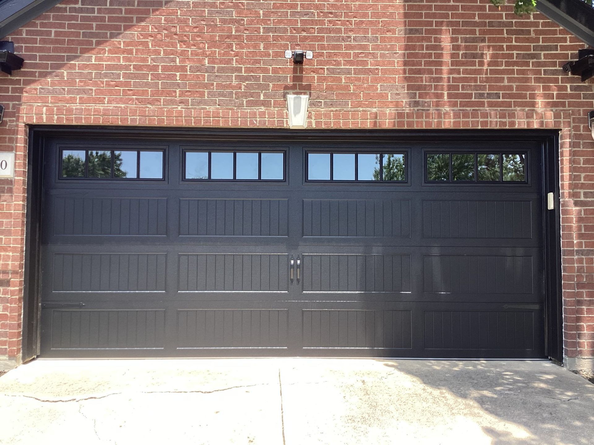 A black residential garage door with a row of window panes at the top, set into a red brick house wall.