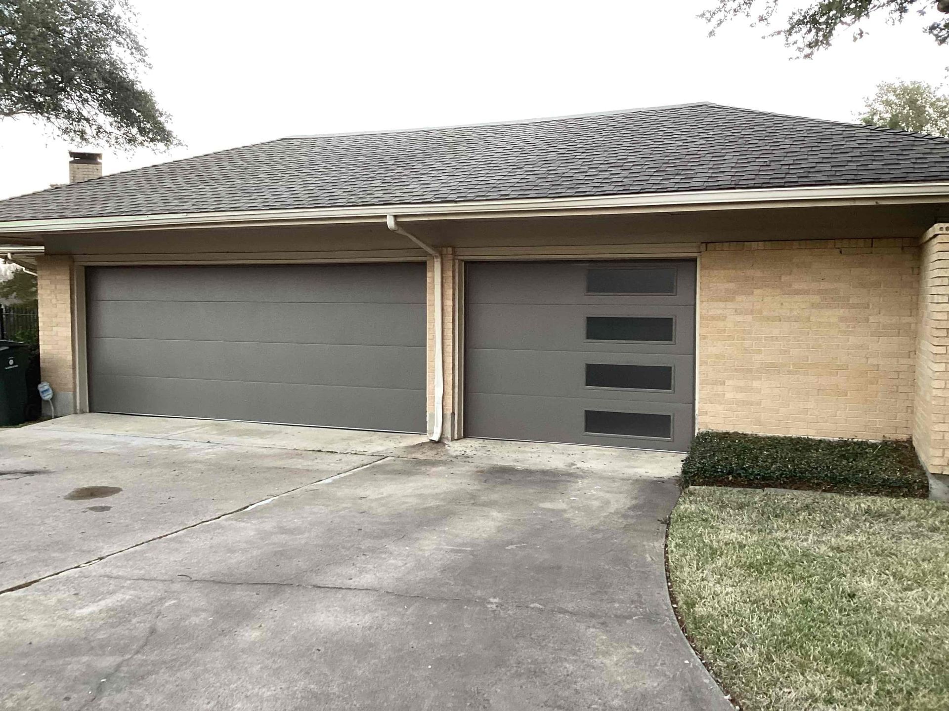 A two-car garage with a gray flat-panel door and a matching door with vertical windows on a house with brick siding.
