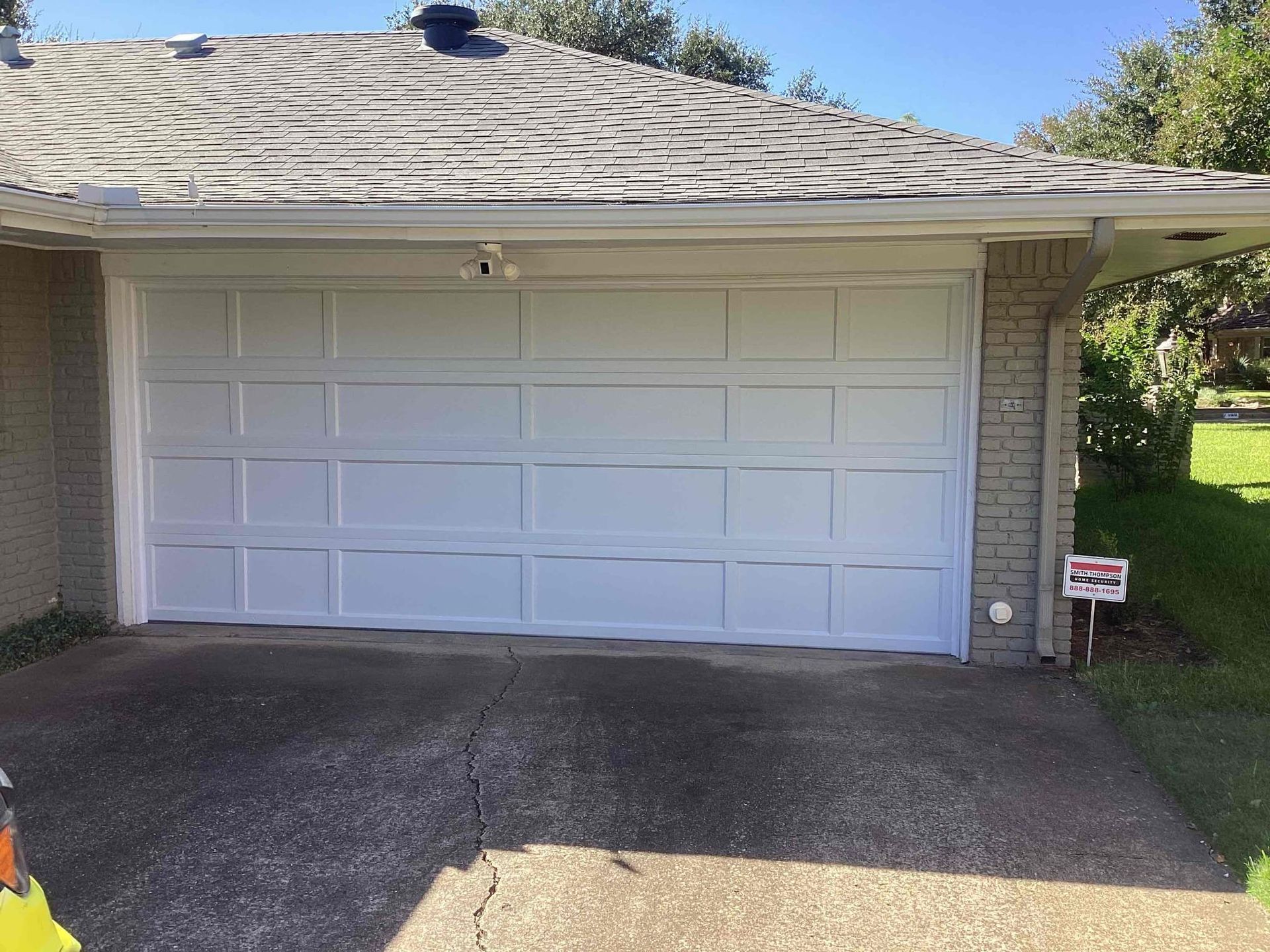 A white, multi-panel garage door on a house with a brick exterior, shingled roof, and a paved driveway.