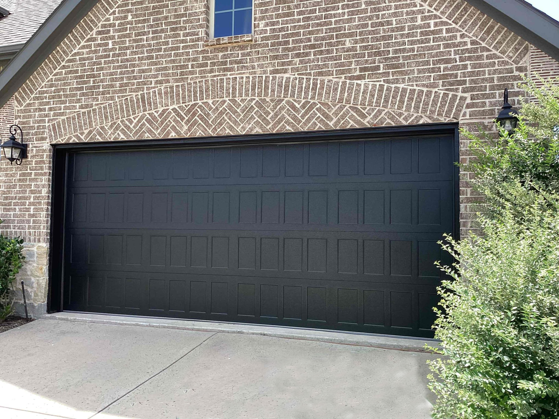 A modern black garage door with rectangular panel detailing, set against a tan brick wall with a small window above.