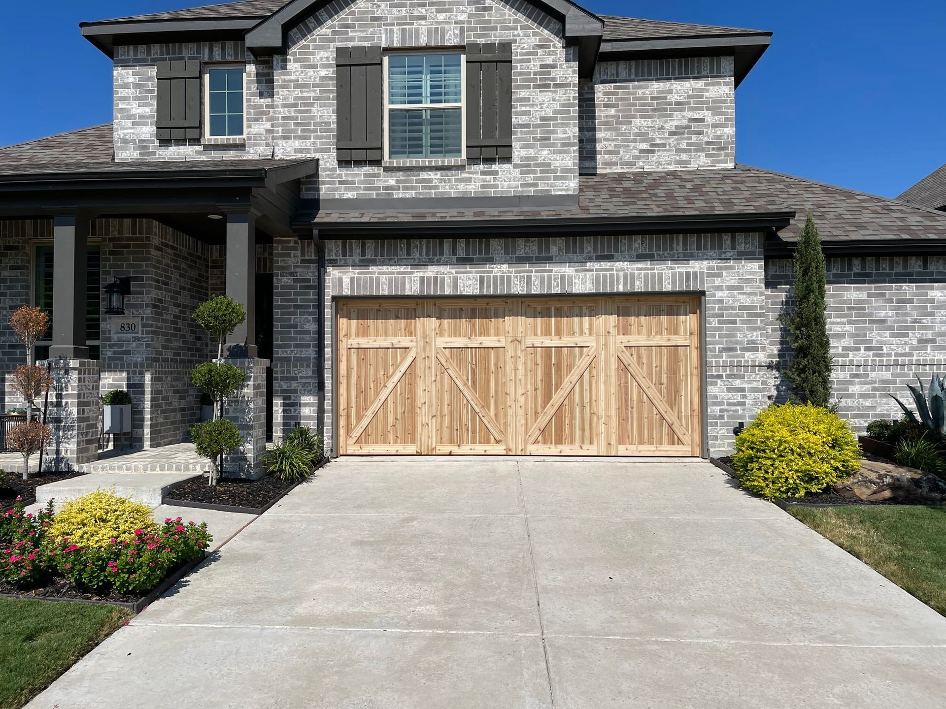 A two-story gray brick house with a wooden garage door and landscaped front yard under a clear blue sky.