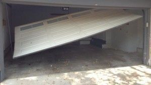 Garage with a white door partially open, appearing damaged and askew. Debris on the concrete floor.