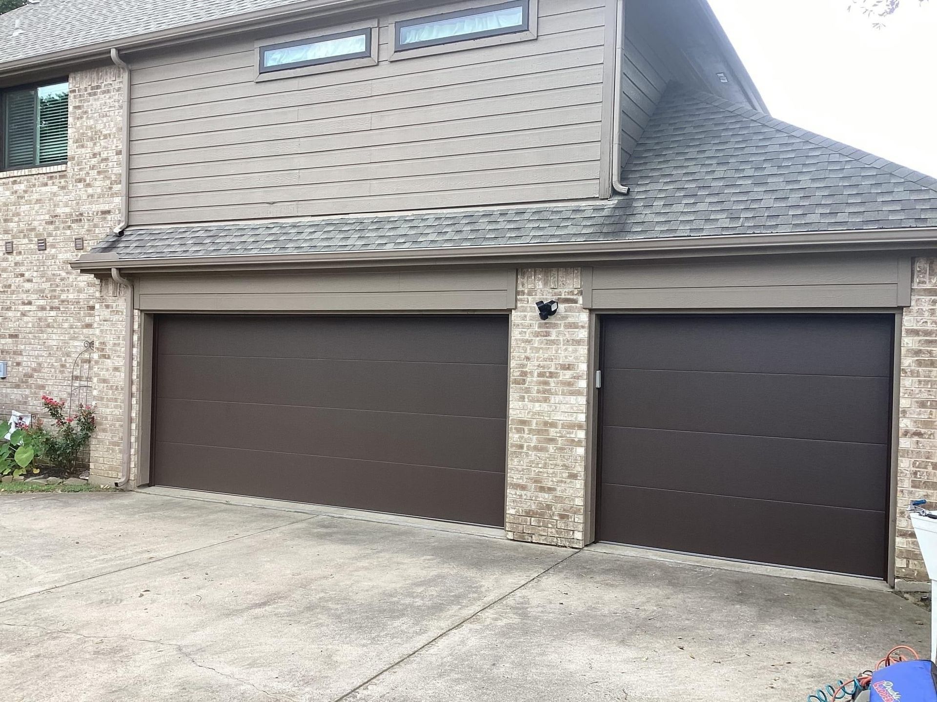 A beige brick house exterior with two modern, dark brown horizontal-panel garage doors and a grey shingled roof.