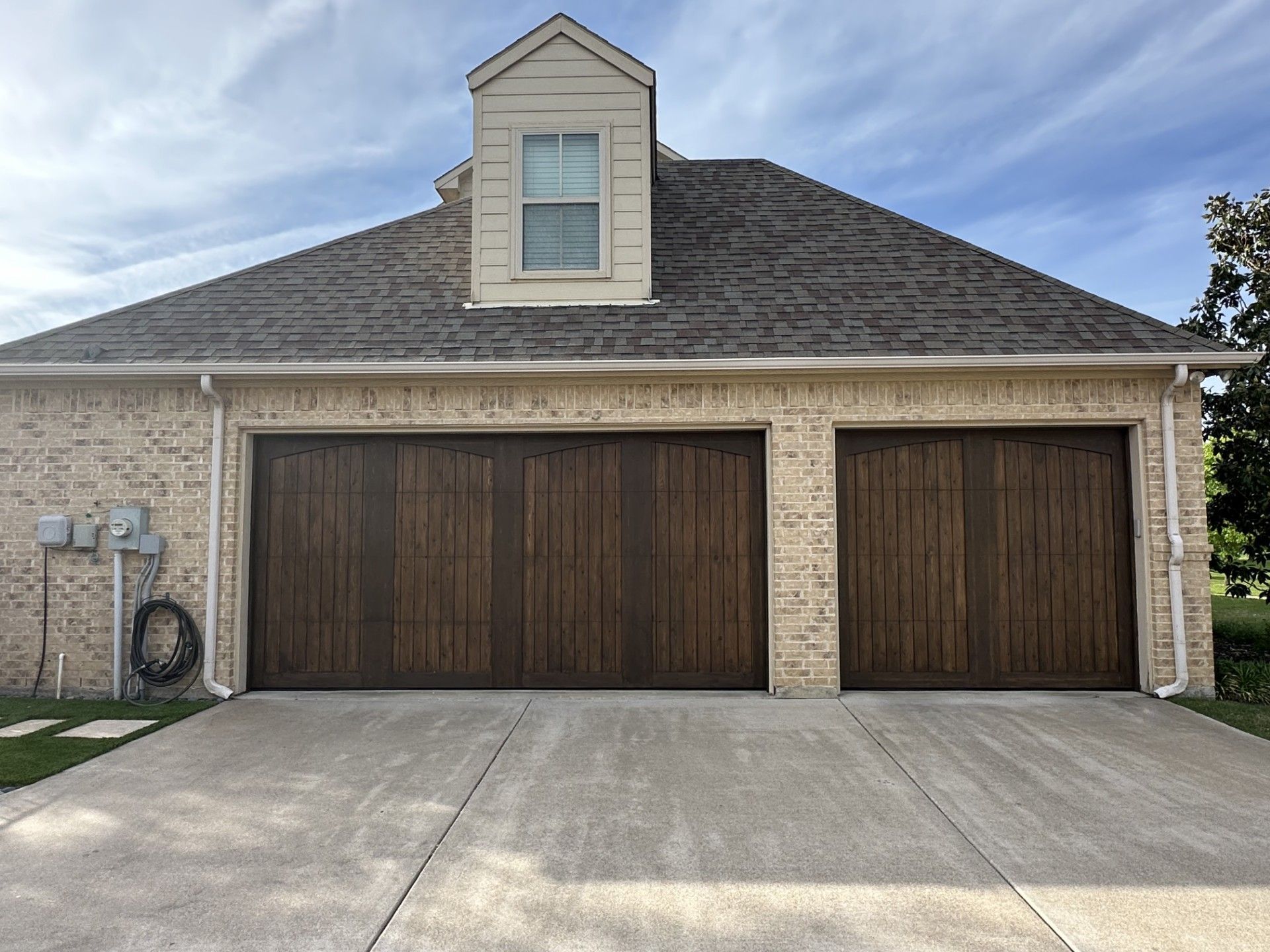 A two-car garage with wood-finished doors, light-colored brick siding, a grey shingled roof, and a central dormer window.