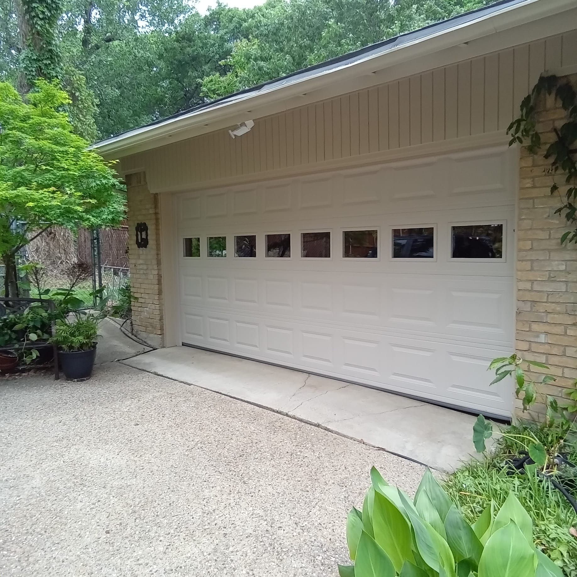Off-white garage door with a row of rectangular windows, attached to a tan brick house with trees and a gravel driveway.