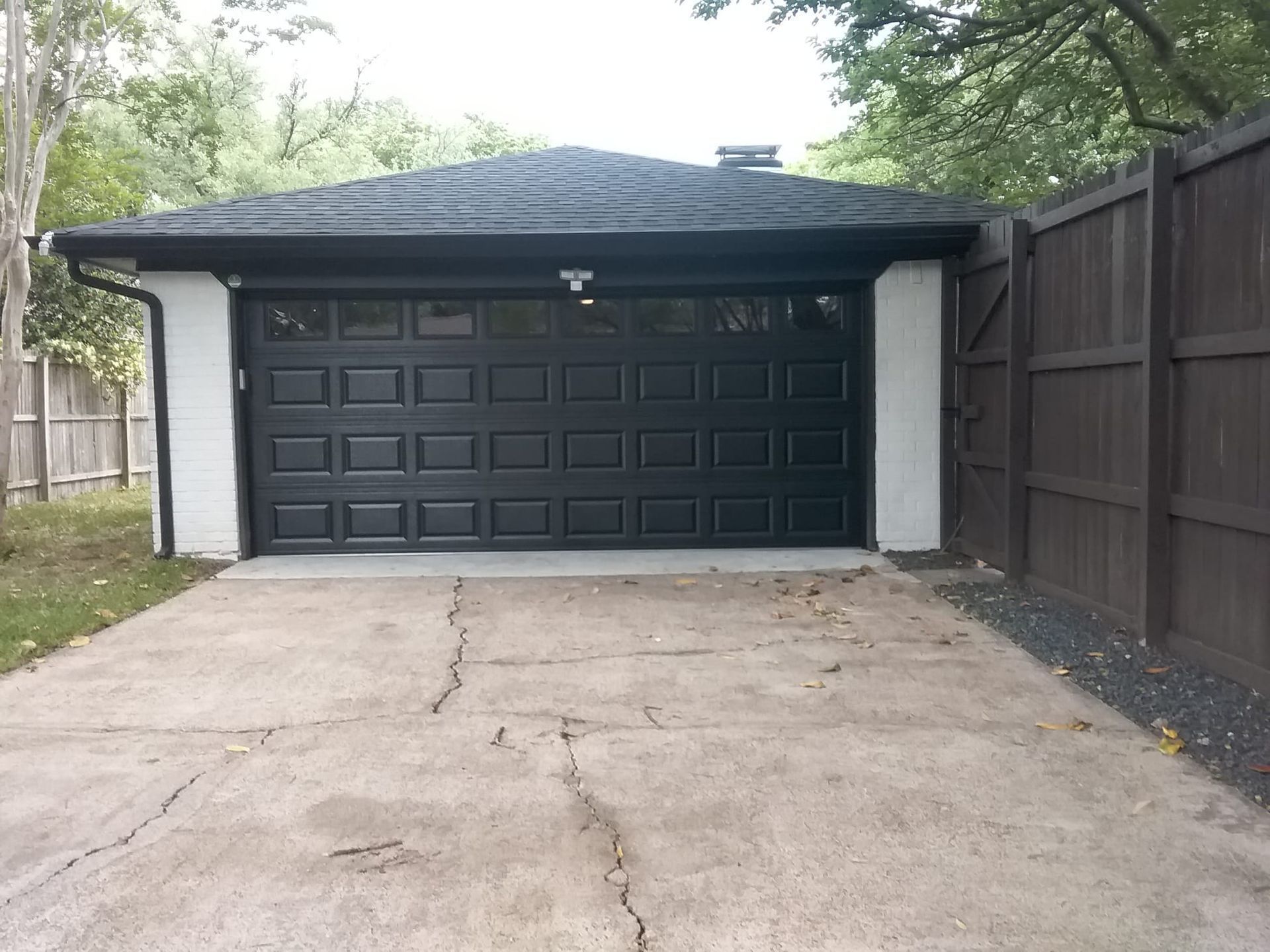 A detached garage with a dark, paneled door and white stucco walls, situated at the end of a cracked concrete driveway.