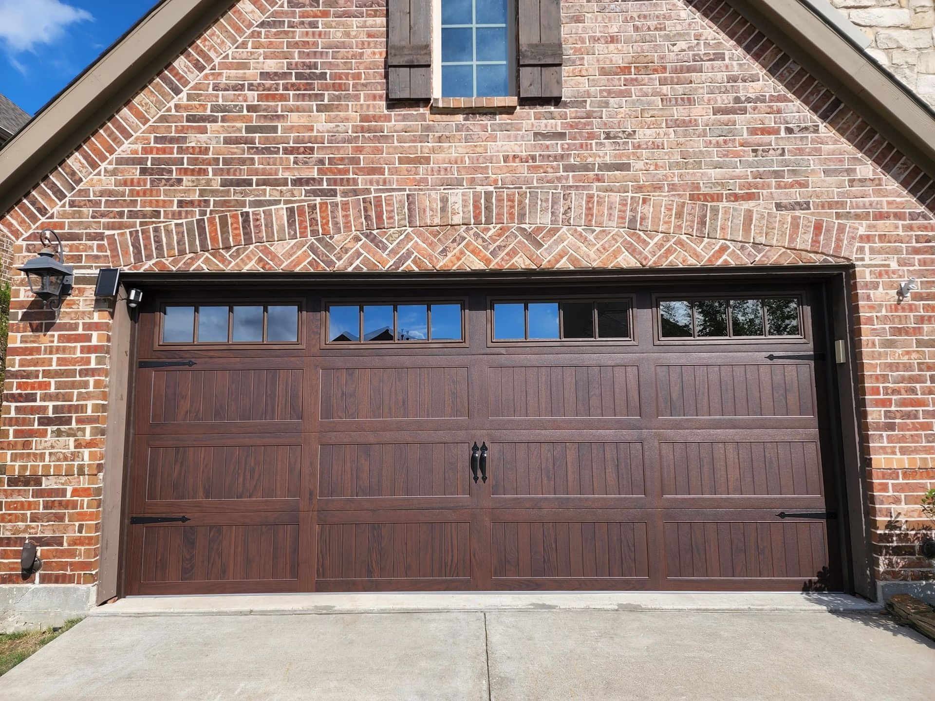 A dark brown, wood-textured garage door with a row of windows sits in a brick home exterior under a gabled roof.