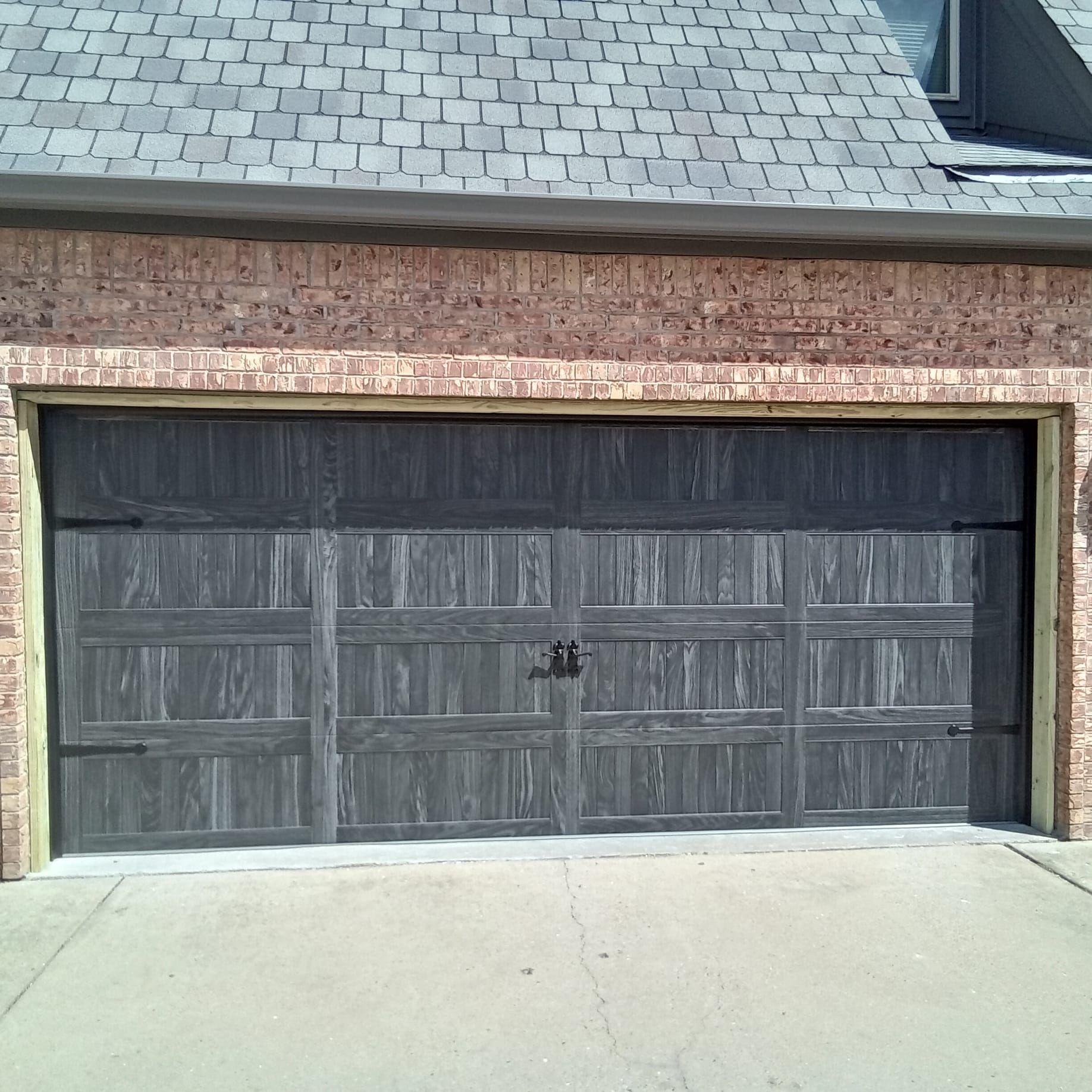 A dark grey, wood-textured, multi-panel garage door with decorative black hinges and a handle, set in a brick frame.