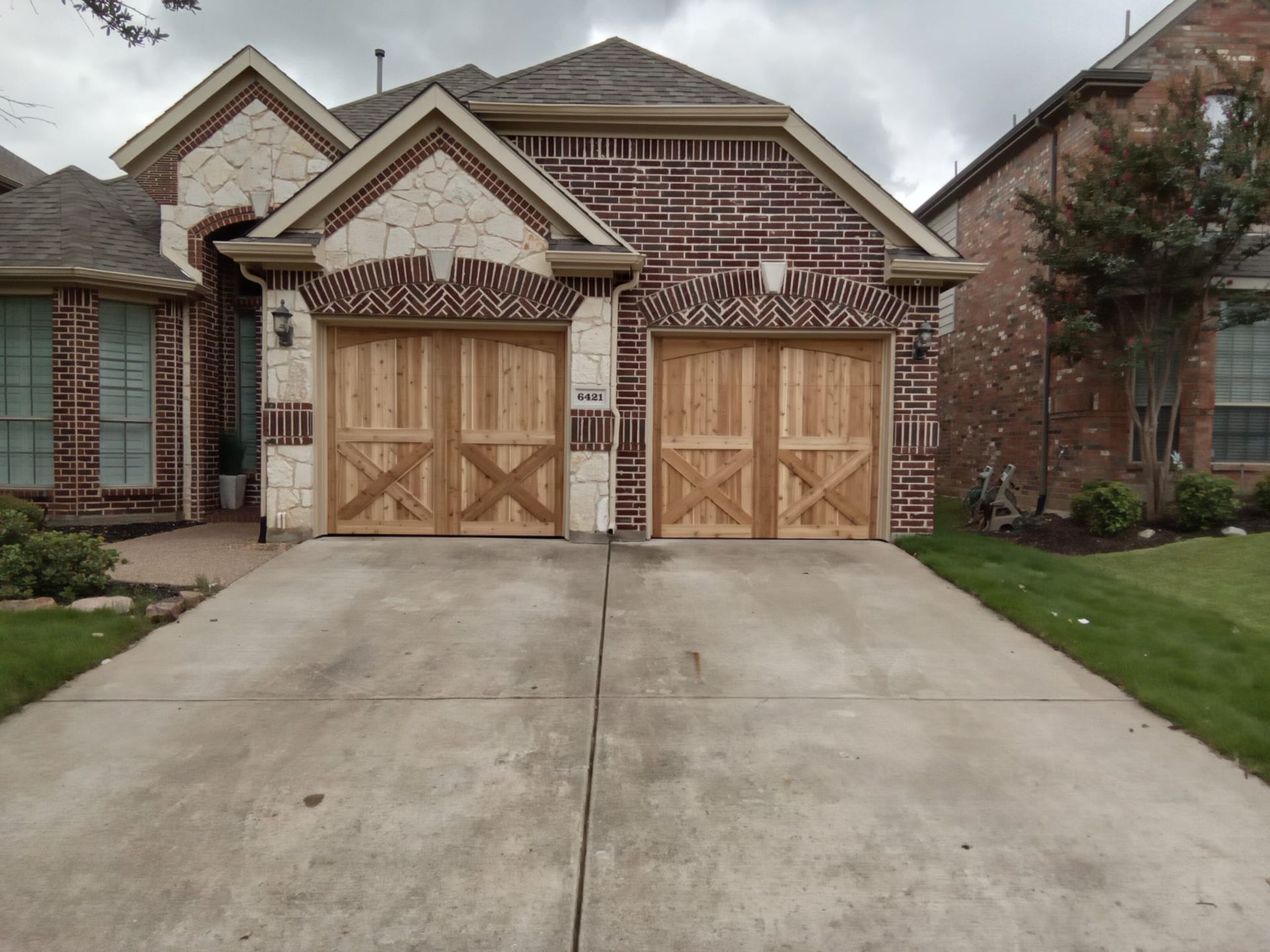 A suburban brick and stone house with two wooden, cross-patterned garage doors and a concrete driveway.
