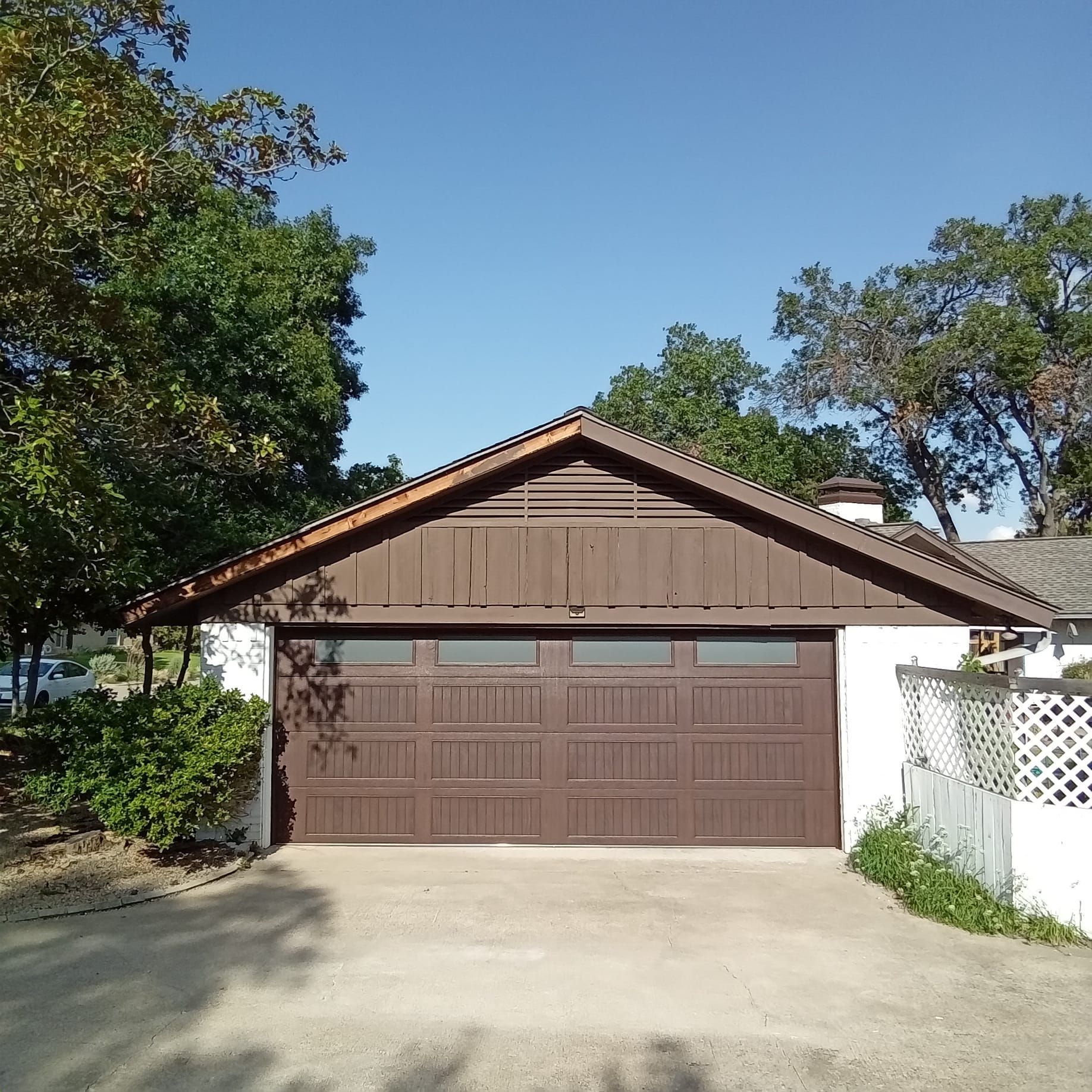 A detached garage with brown siding and a brown door with windows, set in a sunny, tree-lined residential area.
