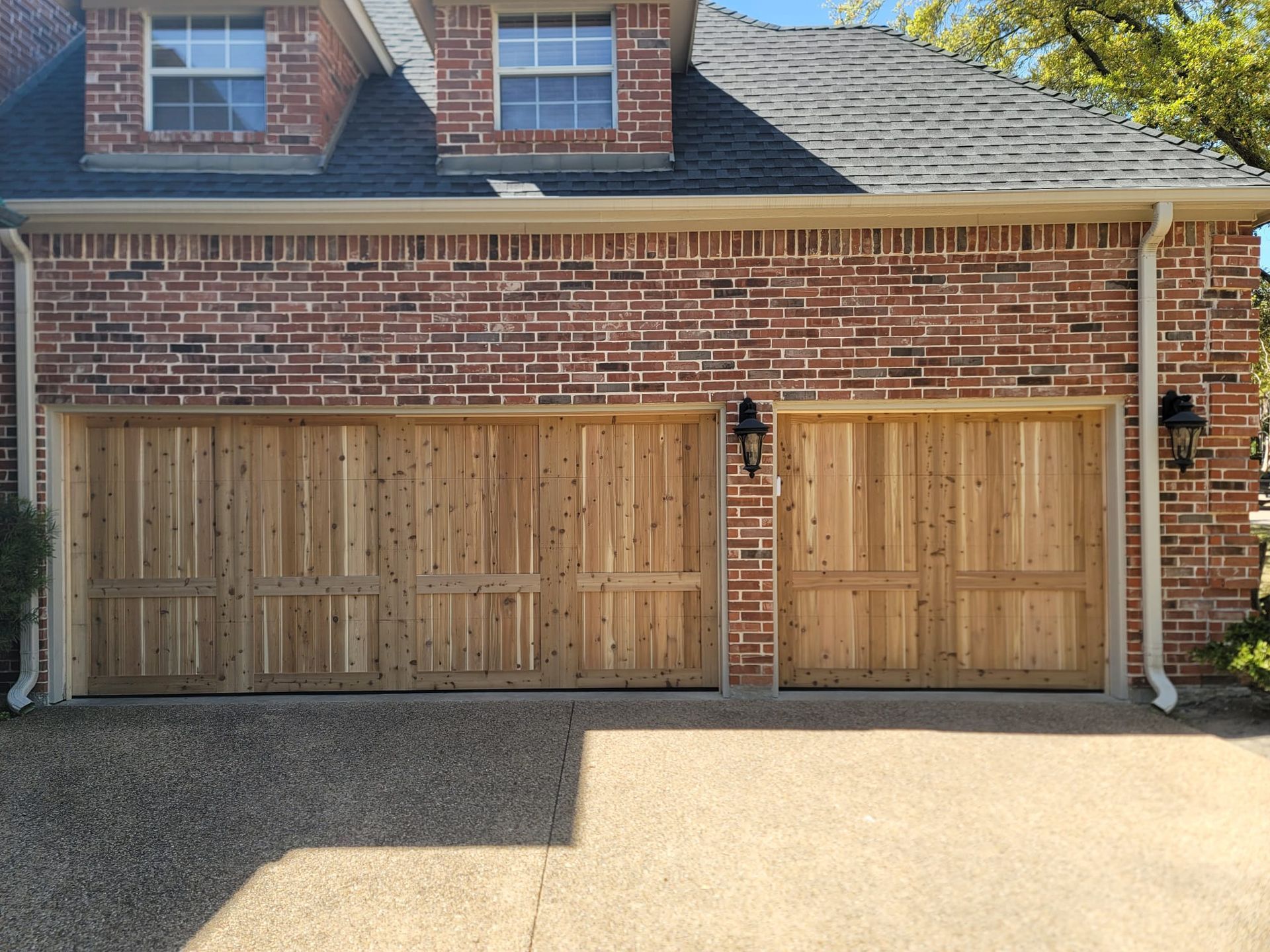 A brick house exterior featuring two natural wood garage doors and two upper dormer windows under a dark shingled roof.