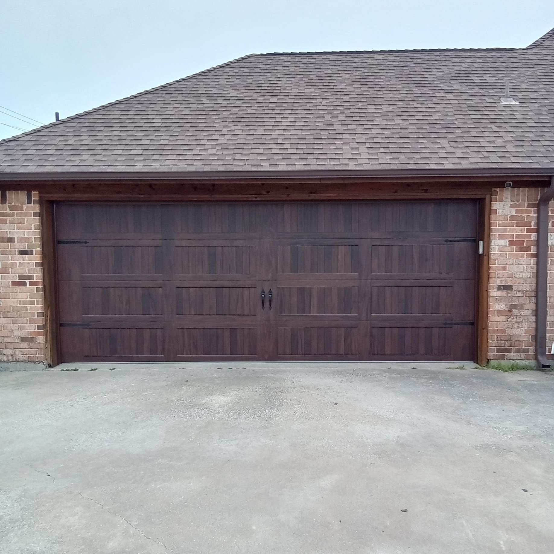 A dark wood-toned garage door with horizontal and vertical paneling, set against a brick wall under a shingled roof.