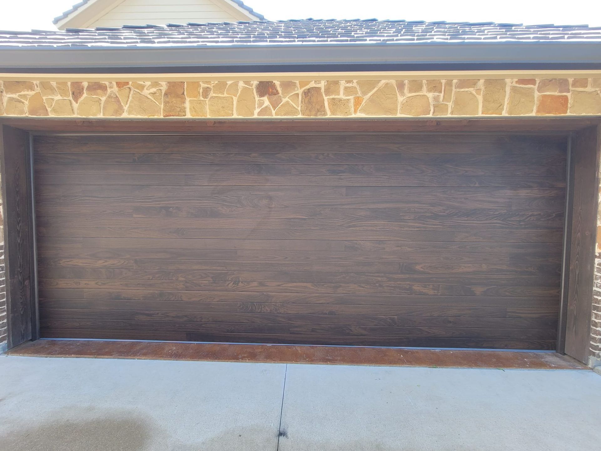 A dark wood-textured garage door framed by a tan stone archway on a concrete driveway.