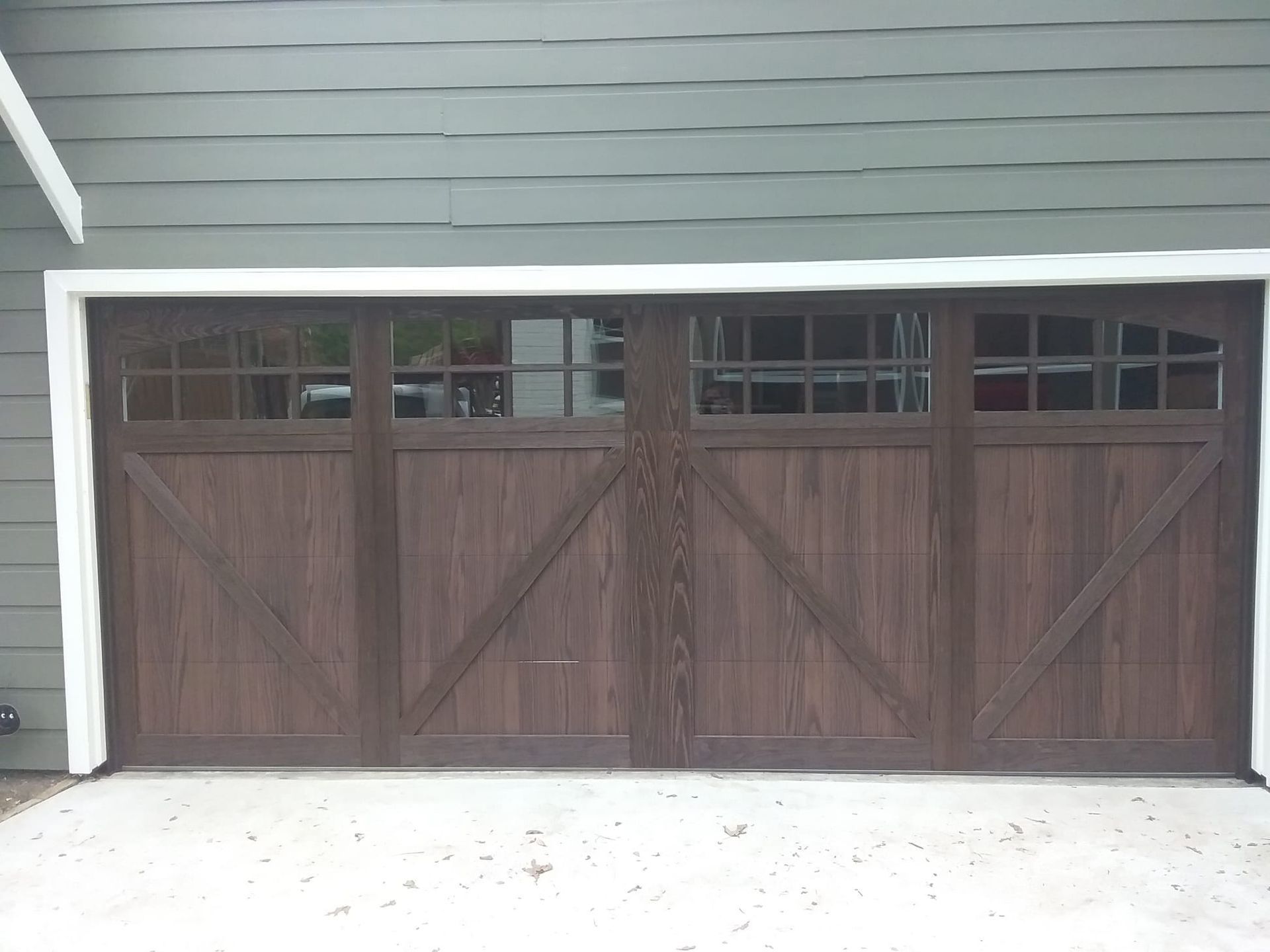 Dark brown, wooden-style garage door with X-bracing and a row of small windows across the top, set against green siding.