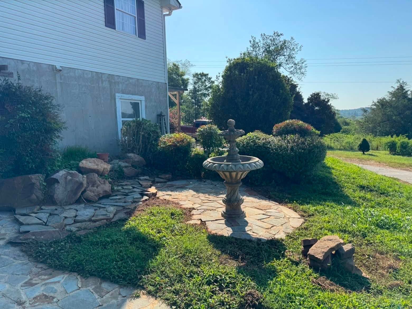 House side yard with stone birdbath, shrubs, rocks, and a grassy path in bright sunlight