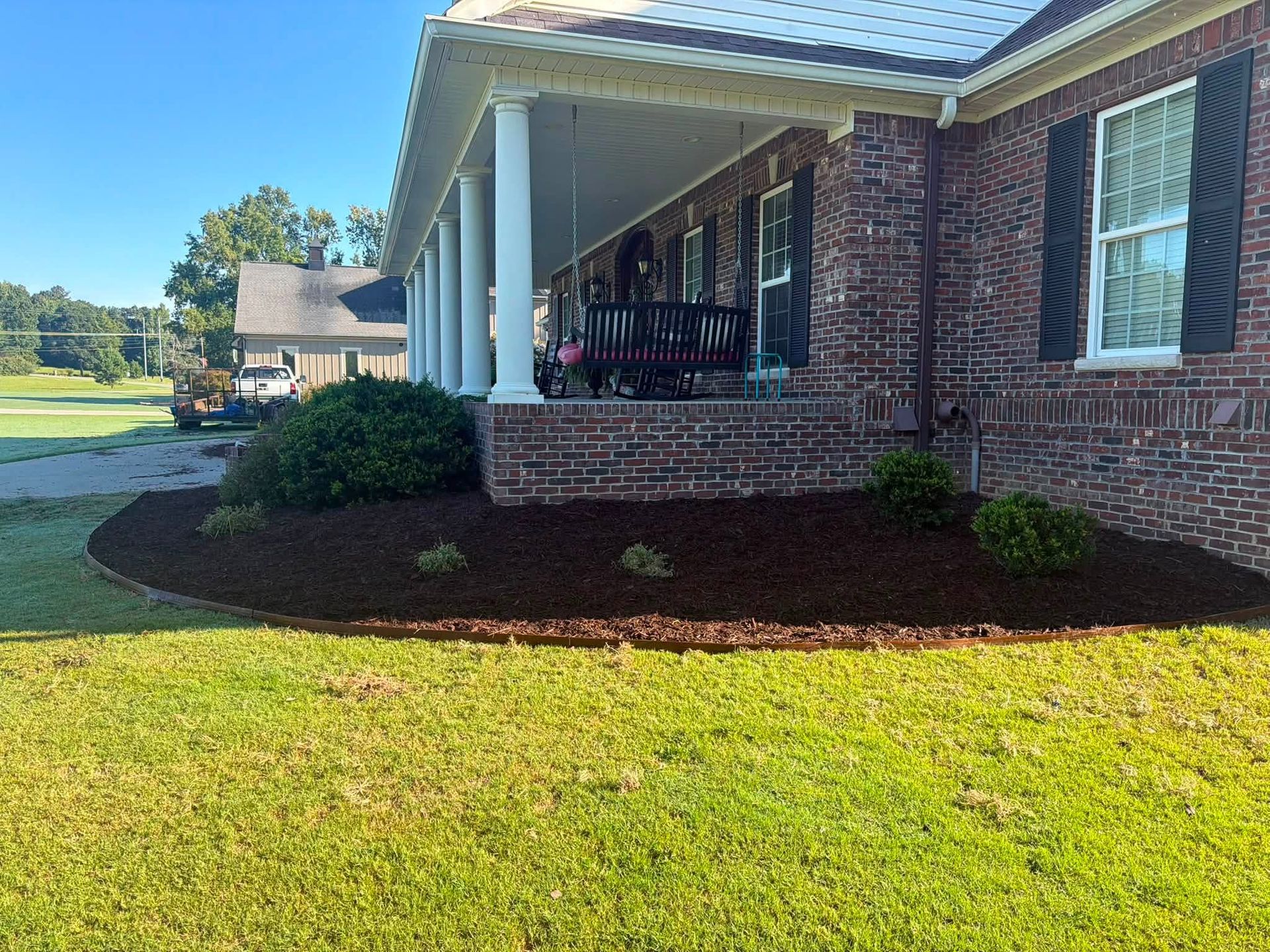 Brick house with white columns, front porch, and landscaped lawn in bright sunlight.