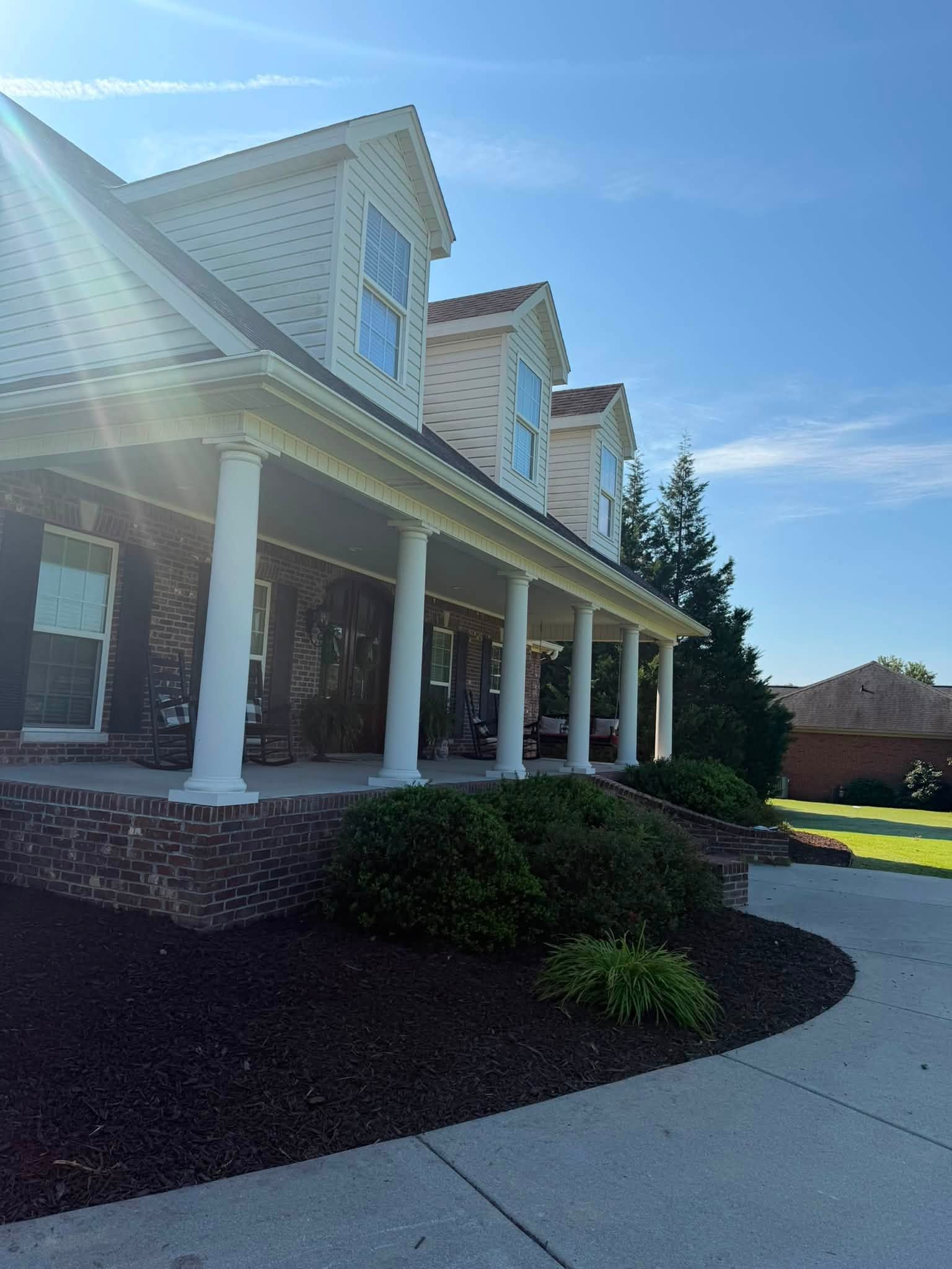 Sunlit suburban house with white porch columns, brick accents, and landscaped front yard under a clear blue sky