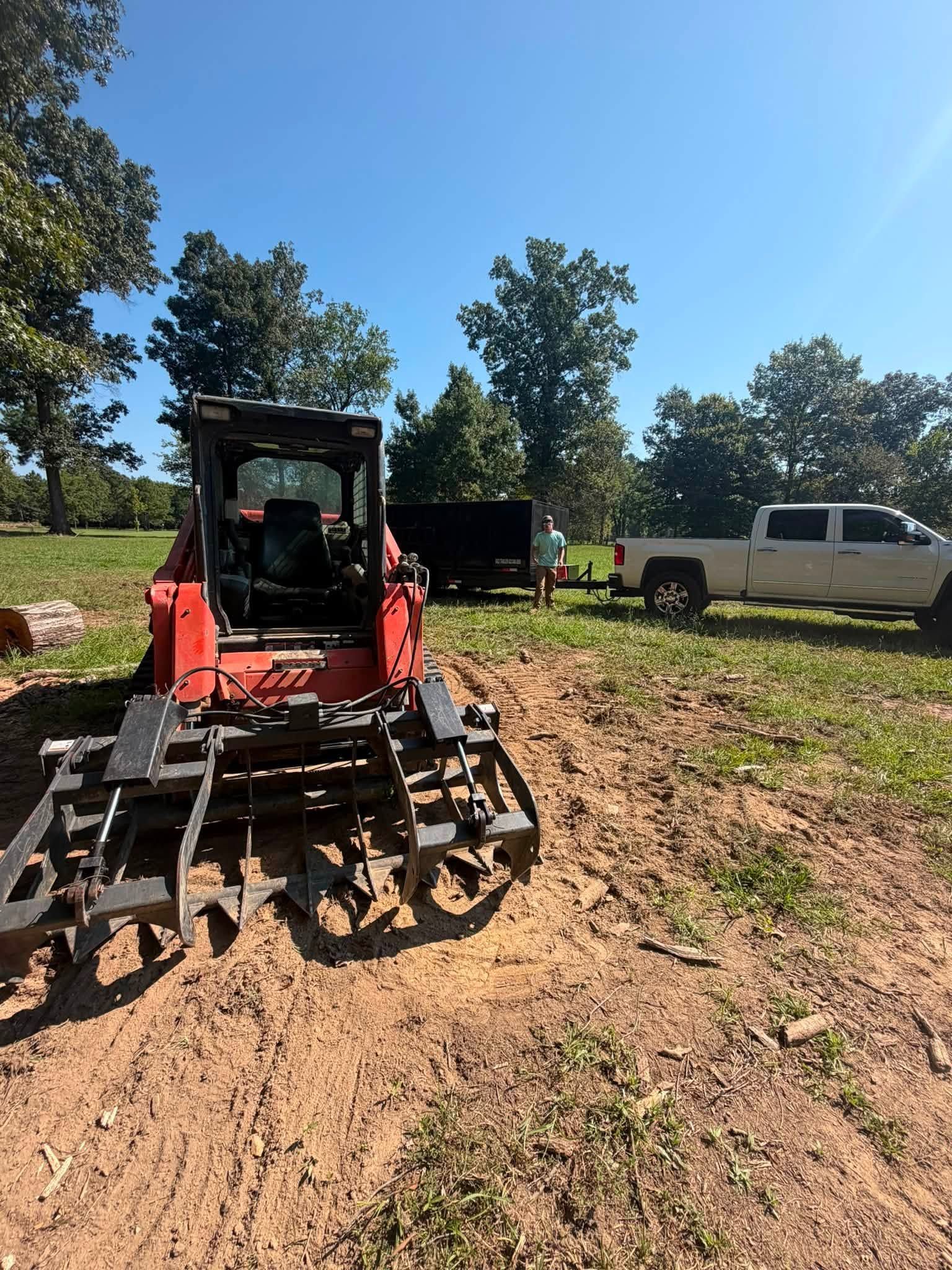 Red skid-steer with grapple on dirt, parked near a white pickup and trees on a sunny day