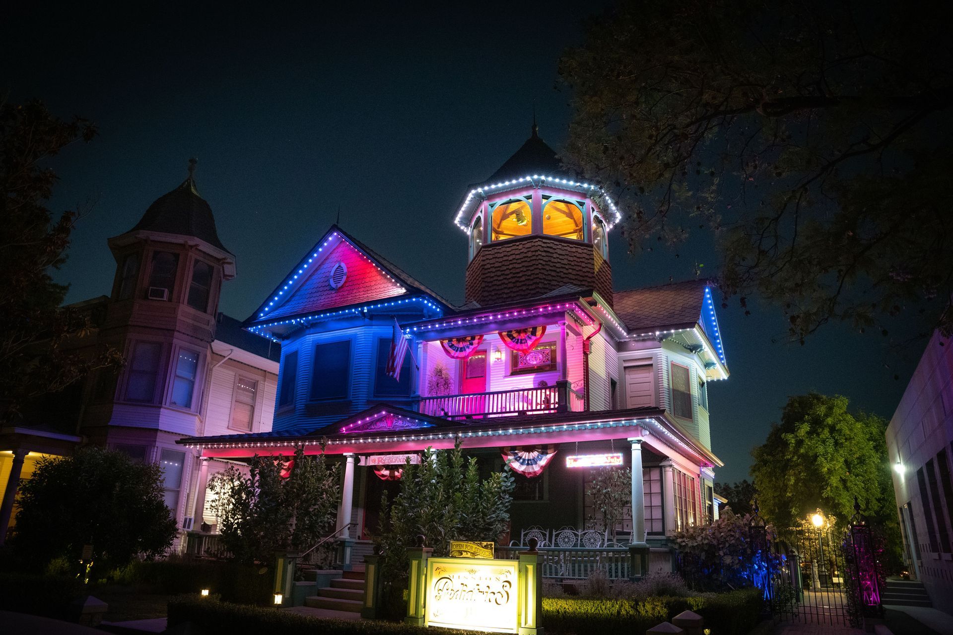 Colorfully lit Victorian house at night with pink, blue, and purple lights and a glowing tower