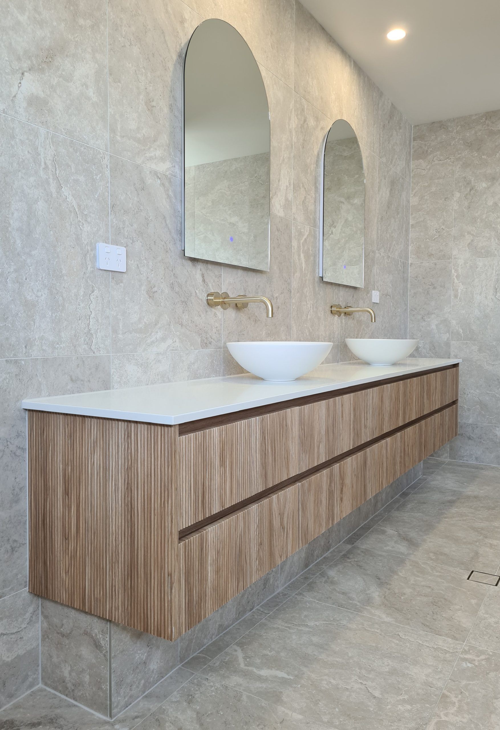 Modern bathroom vanity with two arched mirrors, vessel sinks, and wood-front cabinet against textured tile walls