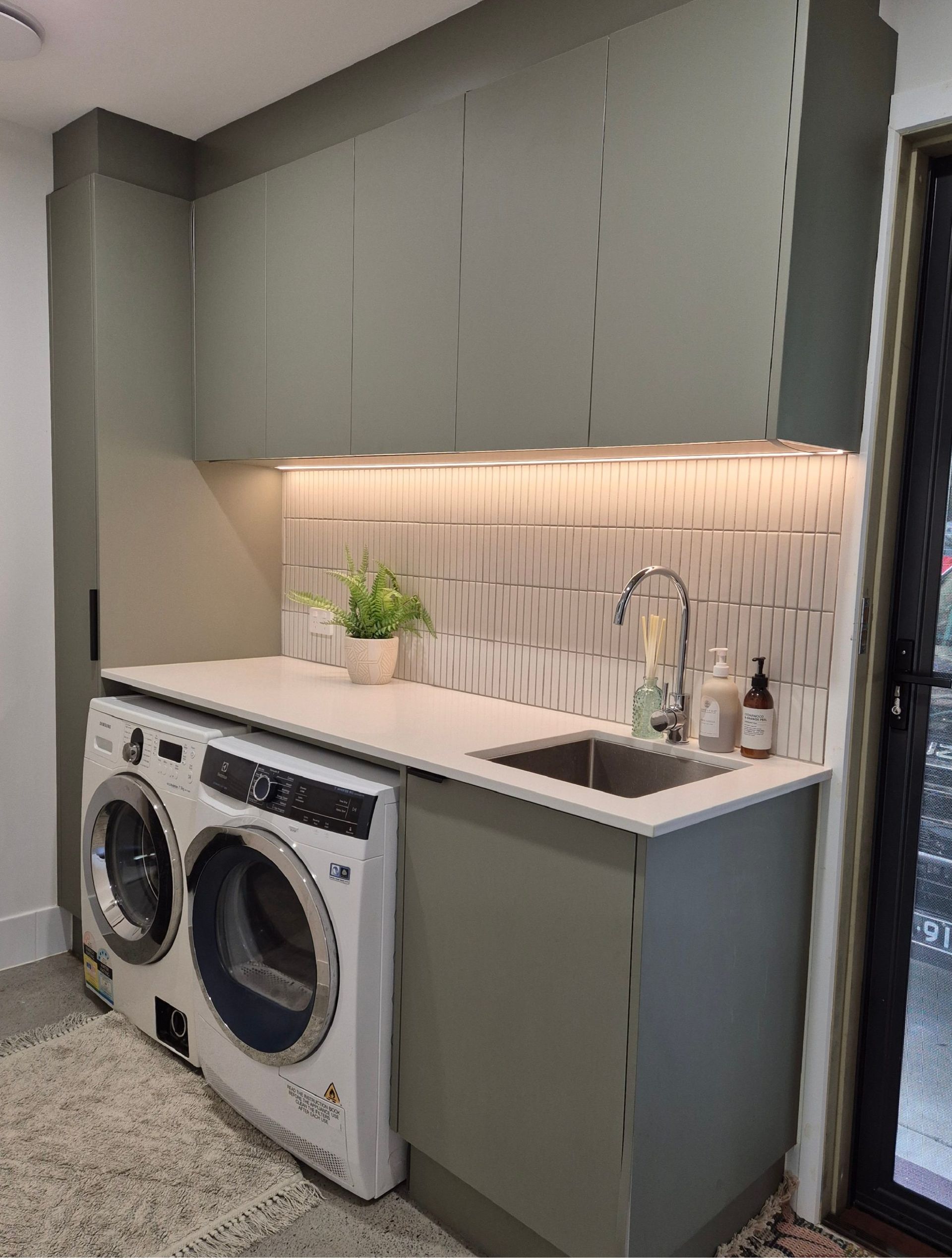 Modern laundry room with stacked washer and dryer, gray cabinets, white countertop, sink, and under-cabinet lighting
