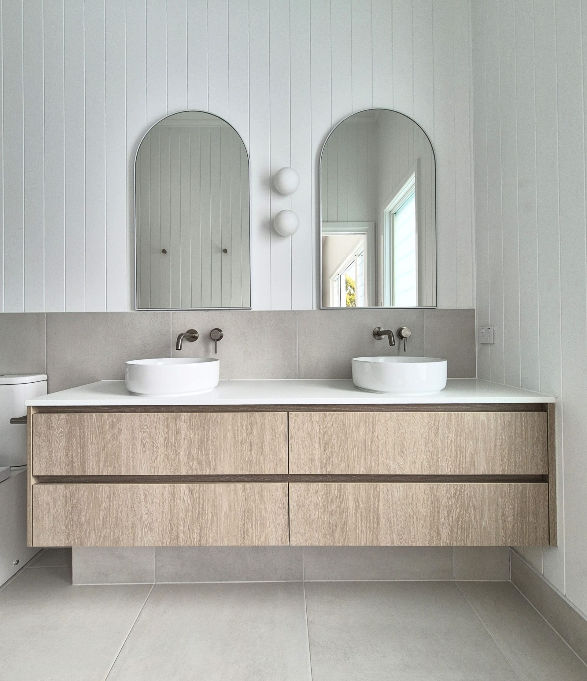 Modern bathroom with dual sinks, arched mirrors, and a light wood vanity against white paneled walls.