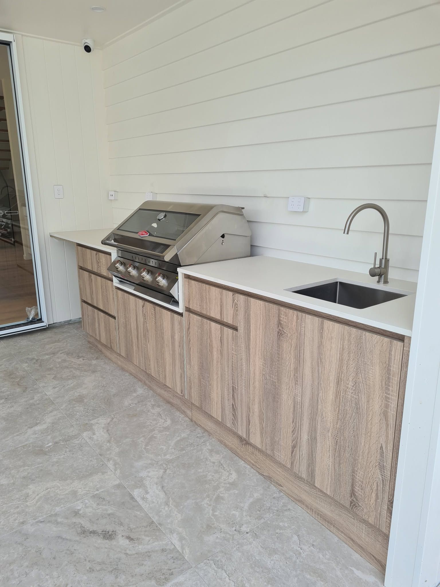 Outdoor kitchen counter with grill, sink, and wood cabinets against a white wall