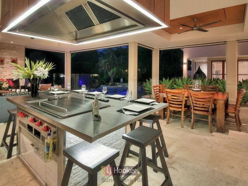 Modern outdoor kitchen with island, bar stools, dining table, and tropical poolside view at night