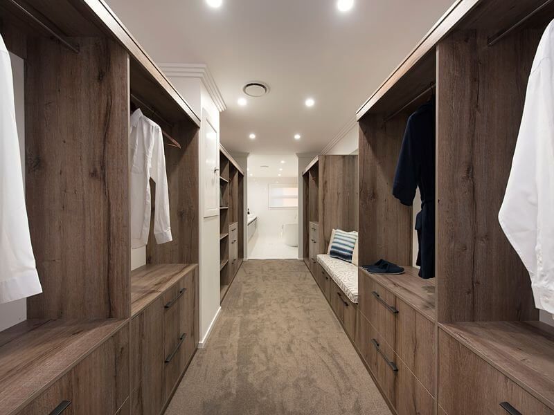 Walk-in closet with wood cabinetry, open shelves, hanging clothes, and a bench at the end of a long aisle.