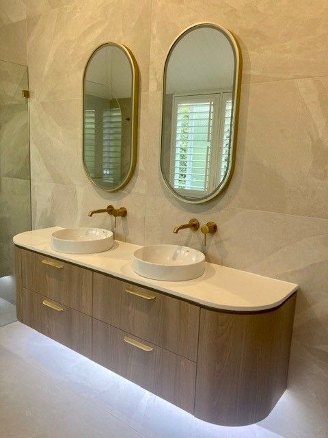 Modern bathroom vanity with two sinks, oval mirrors, and warm wood cabinetry against a stone wall.