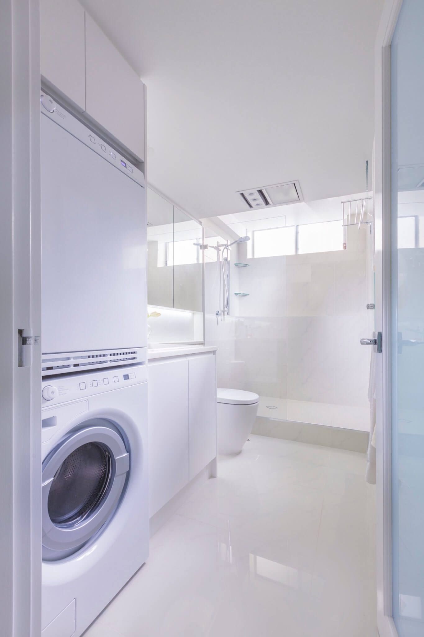 Bright white laundry room with a front-loading washer, tiled floor, and glass door opening to a shower area