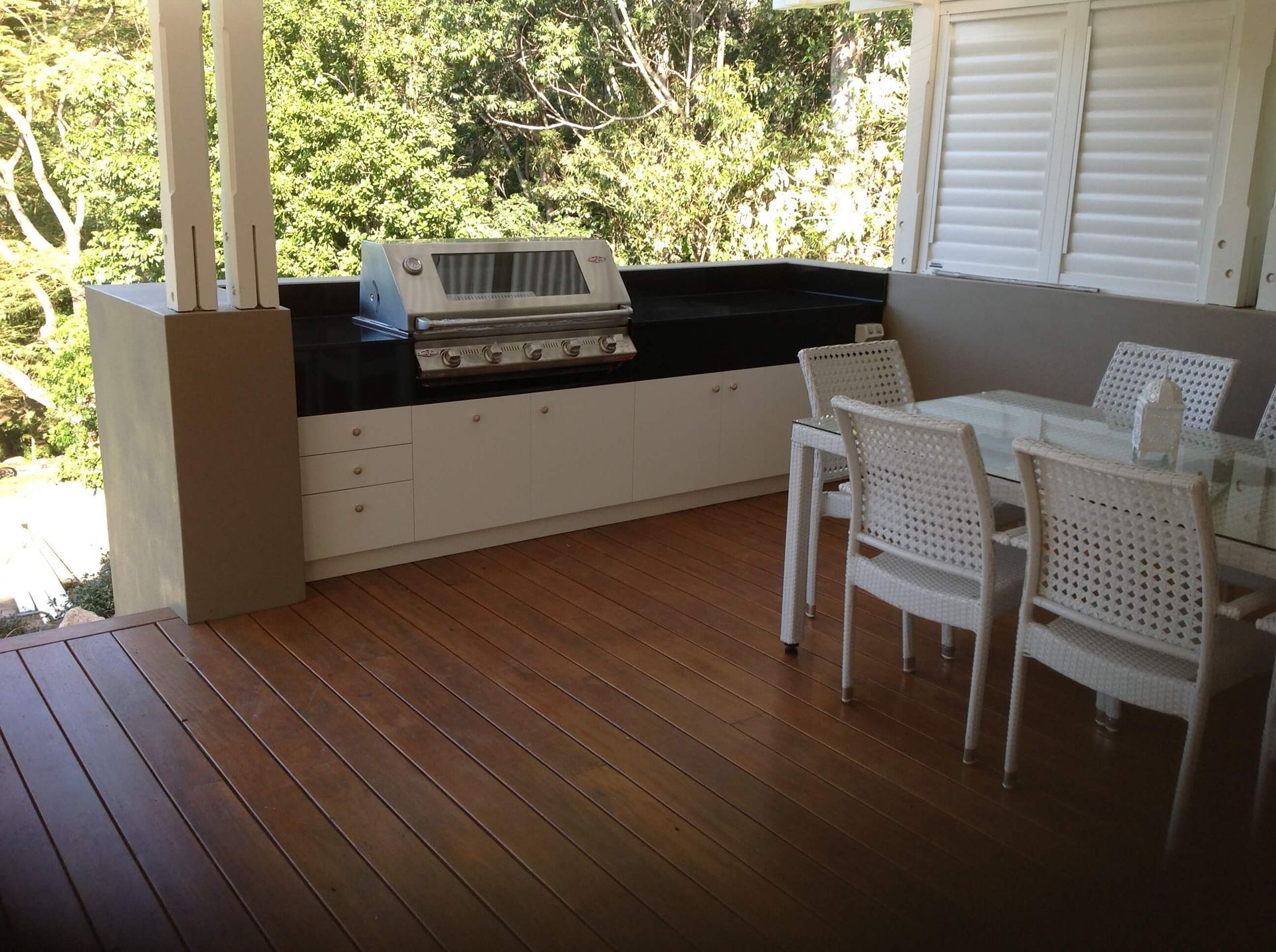 Covered patio with wooden deck, built-in grill, and white dining table and chairs overlooking greenery