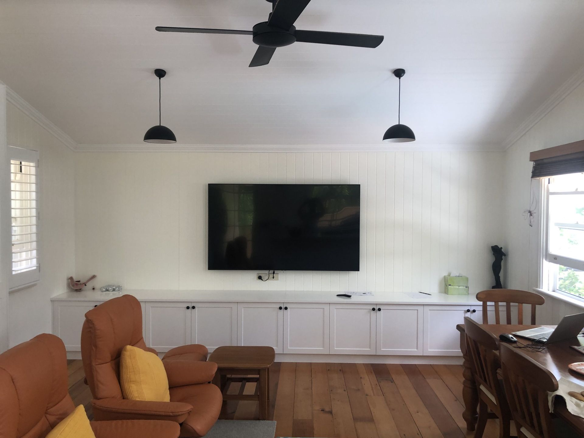 Bright living room with wall-mounted TV, ceiling fan, white shiplap walls, and brown sofa by large windows