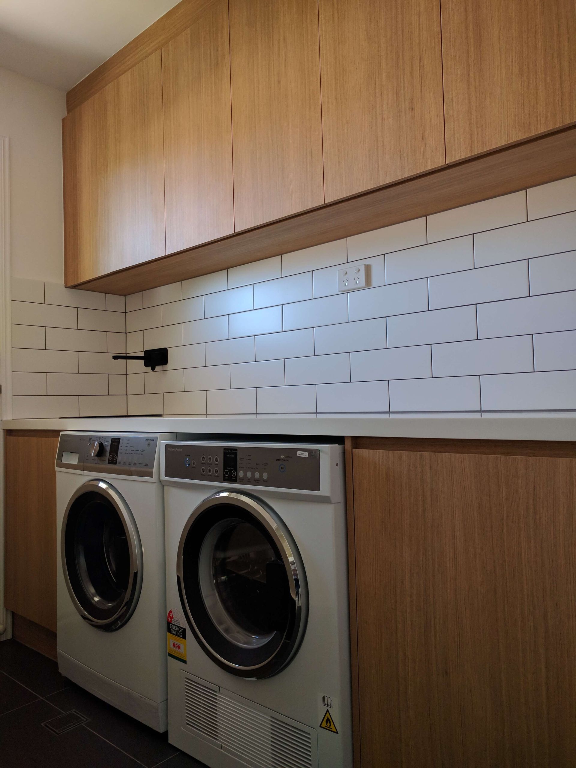 Laundry room with stacked wood cabinets, white tile backsplash, and front-loading washer and dryer underneath.