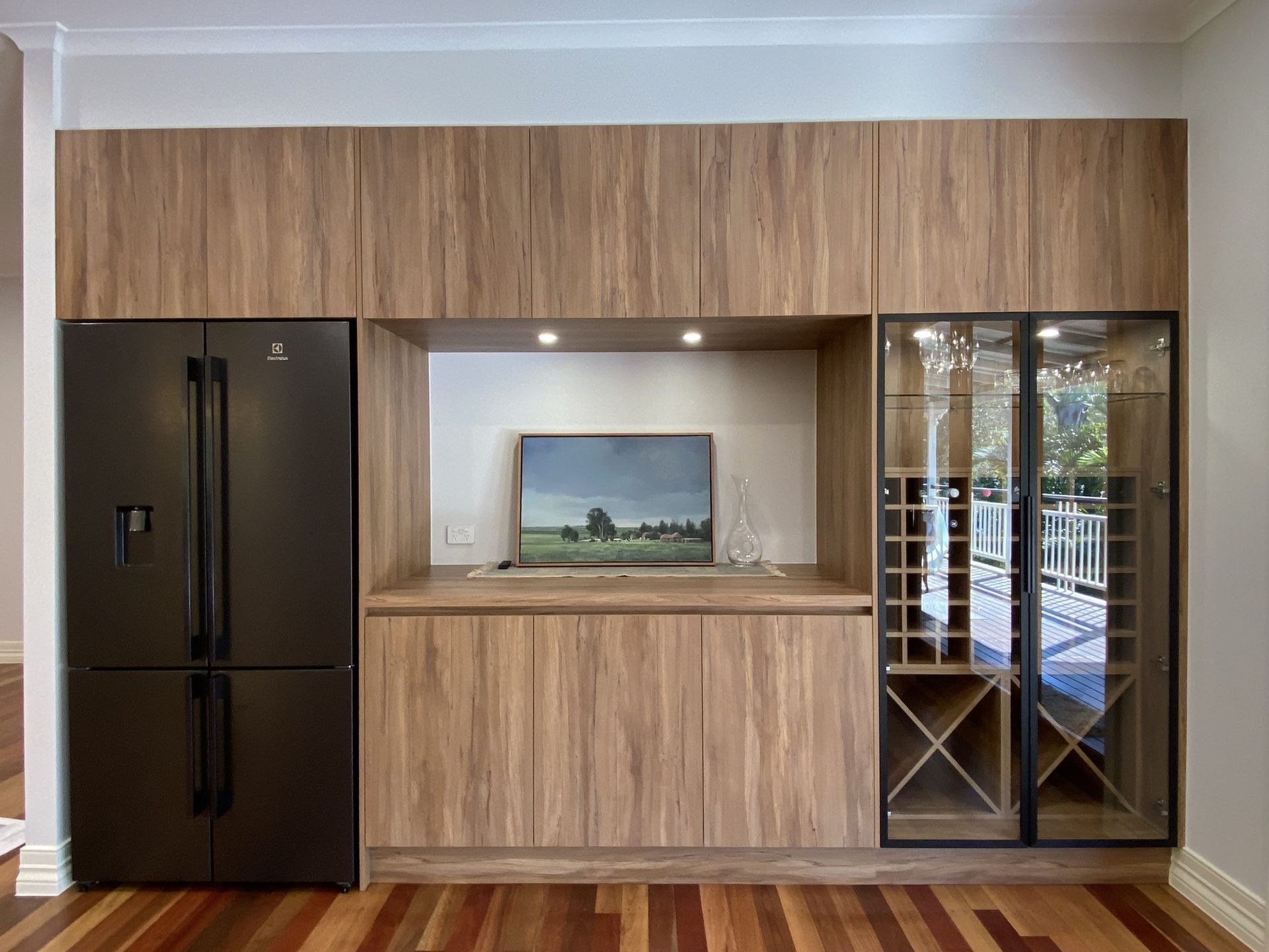 Modern kitchen wall with black refrigerator, wood cabinetry, and mirrored glass cabinet on the right