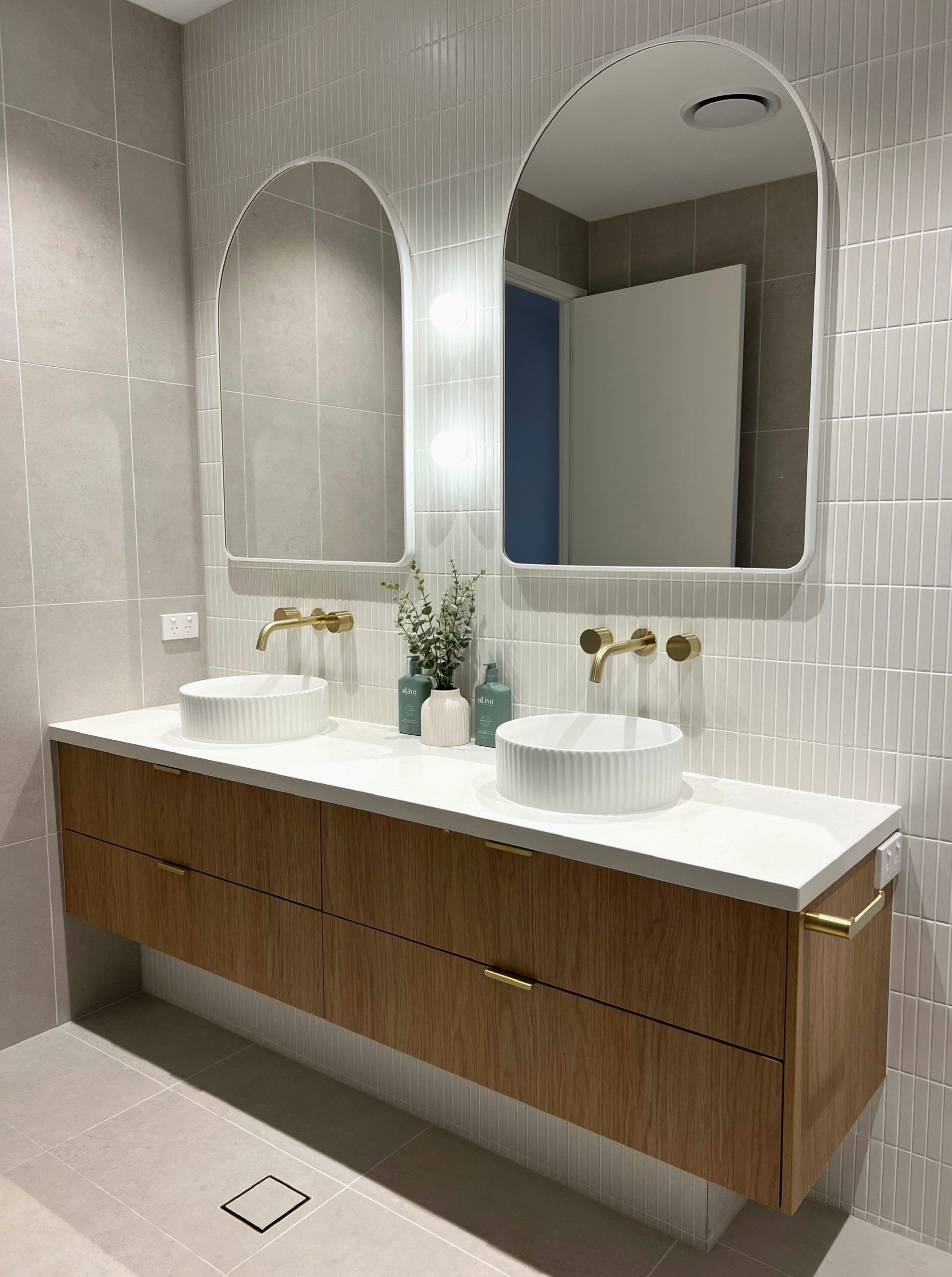 Modern restroom vanity with two vessel sinks, arched mirrors, and wood cabinetry against white tiled walls