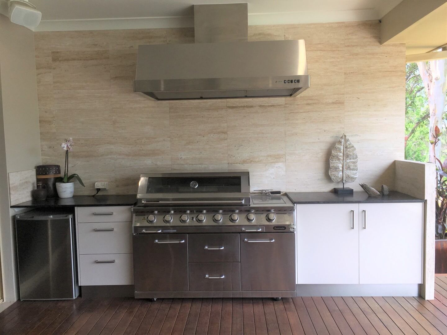 Outdoor kitchen with stainless-steel grill, range hood, sink, and white cabinets under a covered patio