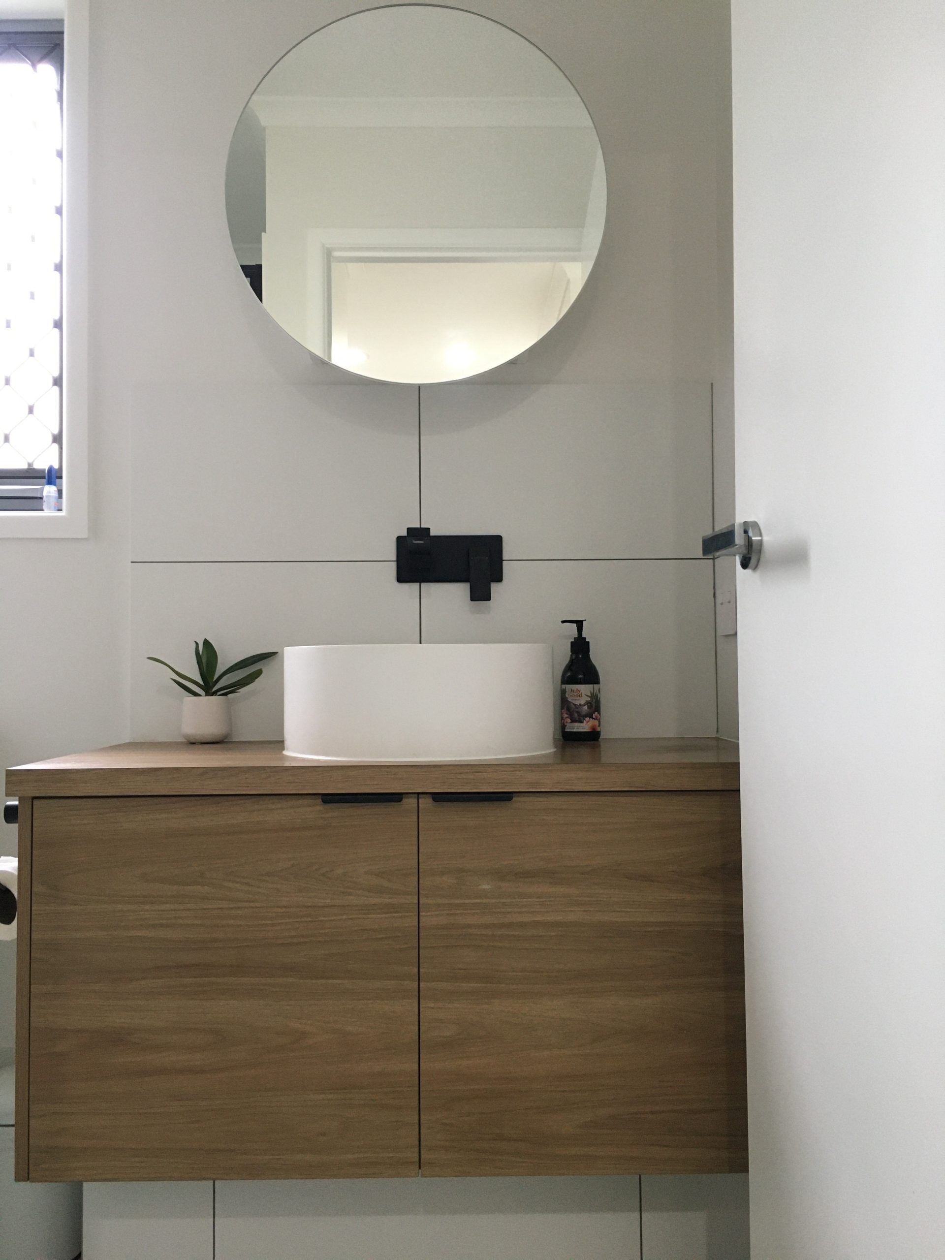 Minimal bathroom vanity with wood cabinet, round mirror, small plant, and soap dispenser against a gray wall.