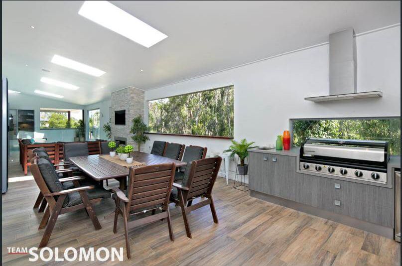 Modern open-plan dining and kitchen area with a long table, wooden chairs, large windows, and built-in grill.