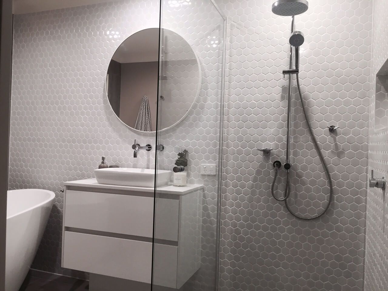 Modern bathroom with white vanity, round mirror, bathtub edge, and tiled shower with handheld sprayer.