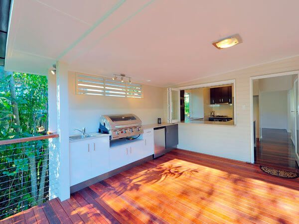 Covered patio with built-in grill, sink, and white cabinetry, opening to a wooded view with warm sunlight on the floor.