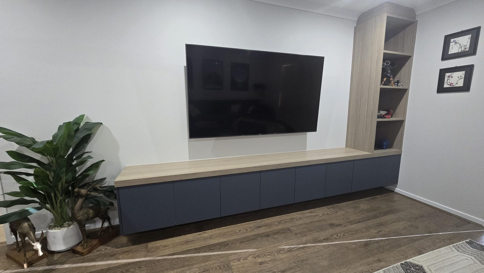 Modern living room with wall-mounted TV, long blue cabinet, wood floors, and potted plant.