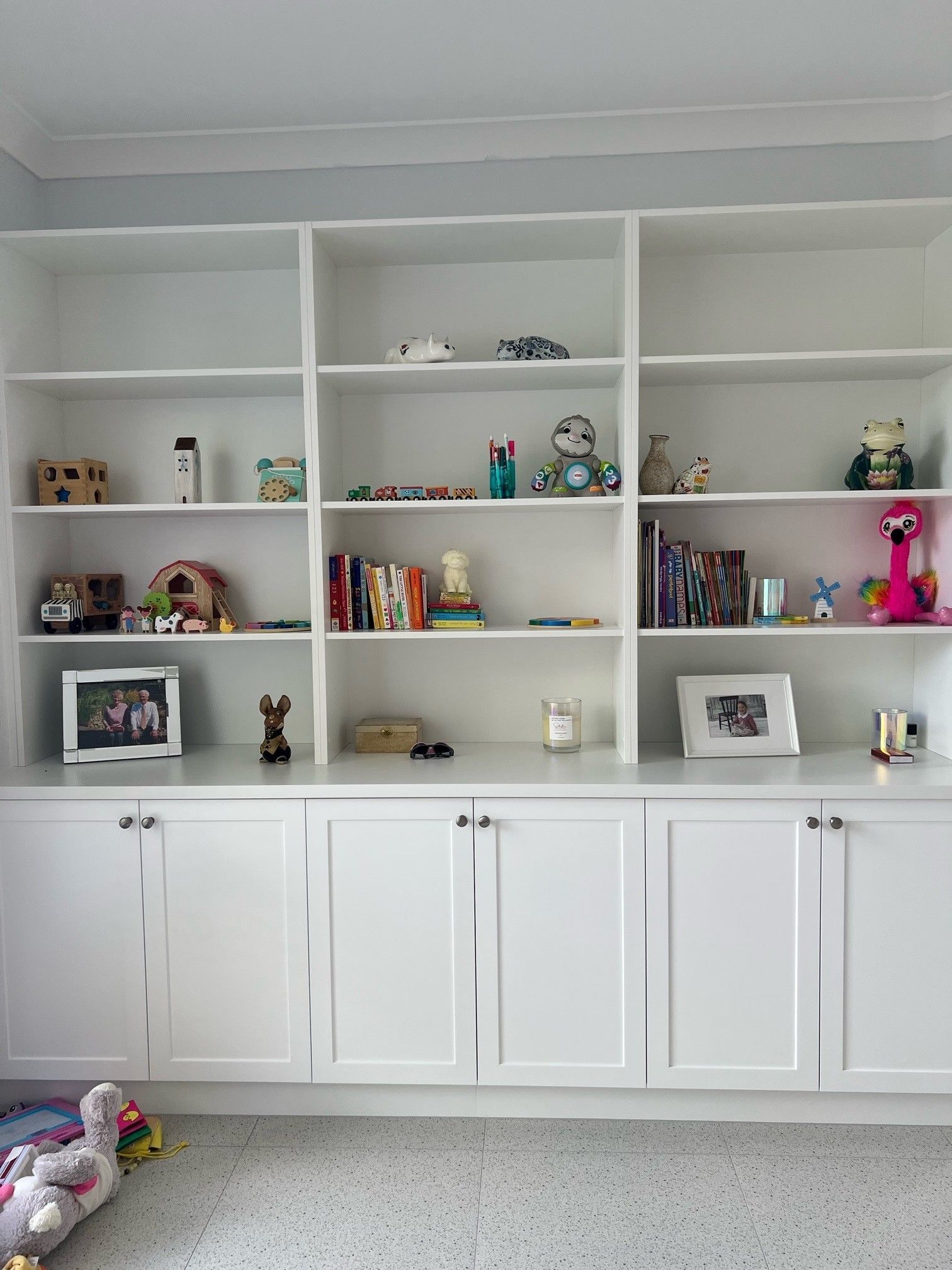 White built-in shelves and cabinets displaying books, toys, and framed photos in a bright room