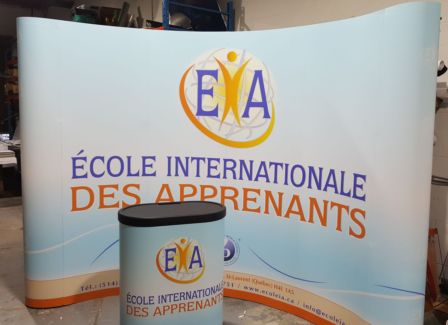 Stand éphémère avec le logo de l'École Internationale des Apprenants en bleu, orange et blanc. Une table cylindrique assortie se trouve devant.