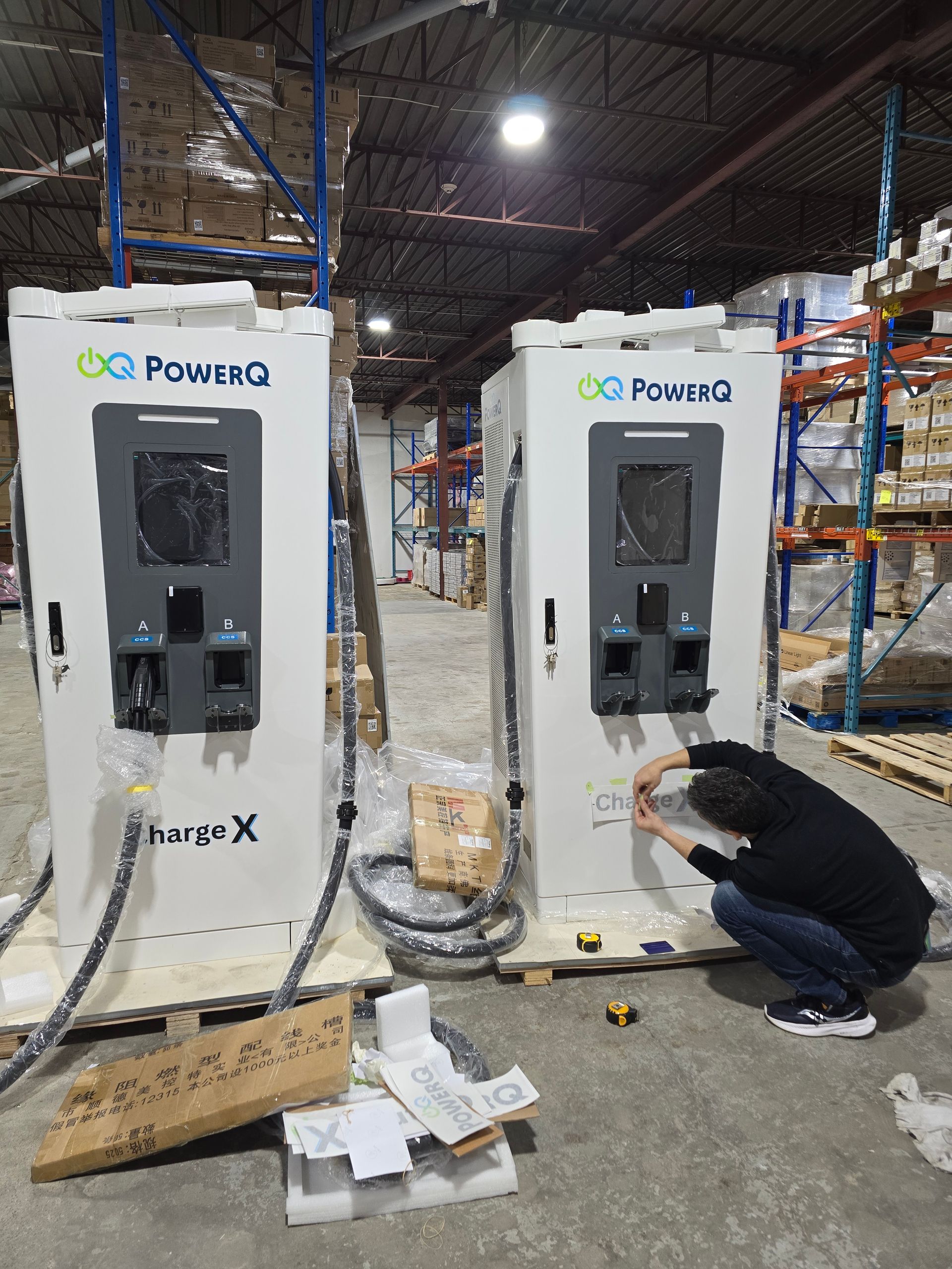 Two white electric vehicle chargers, a person working on one in a warehouse.