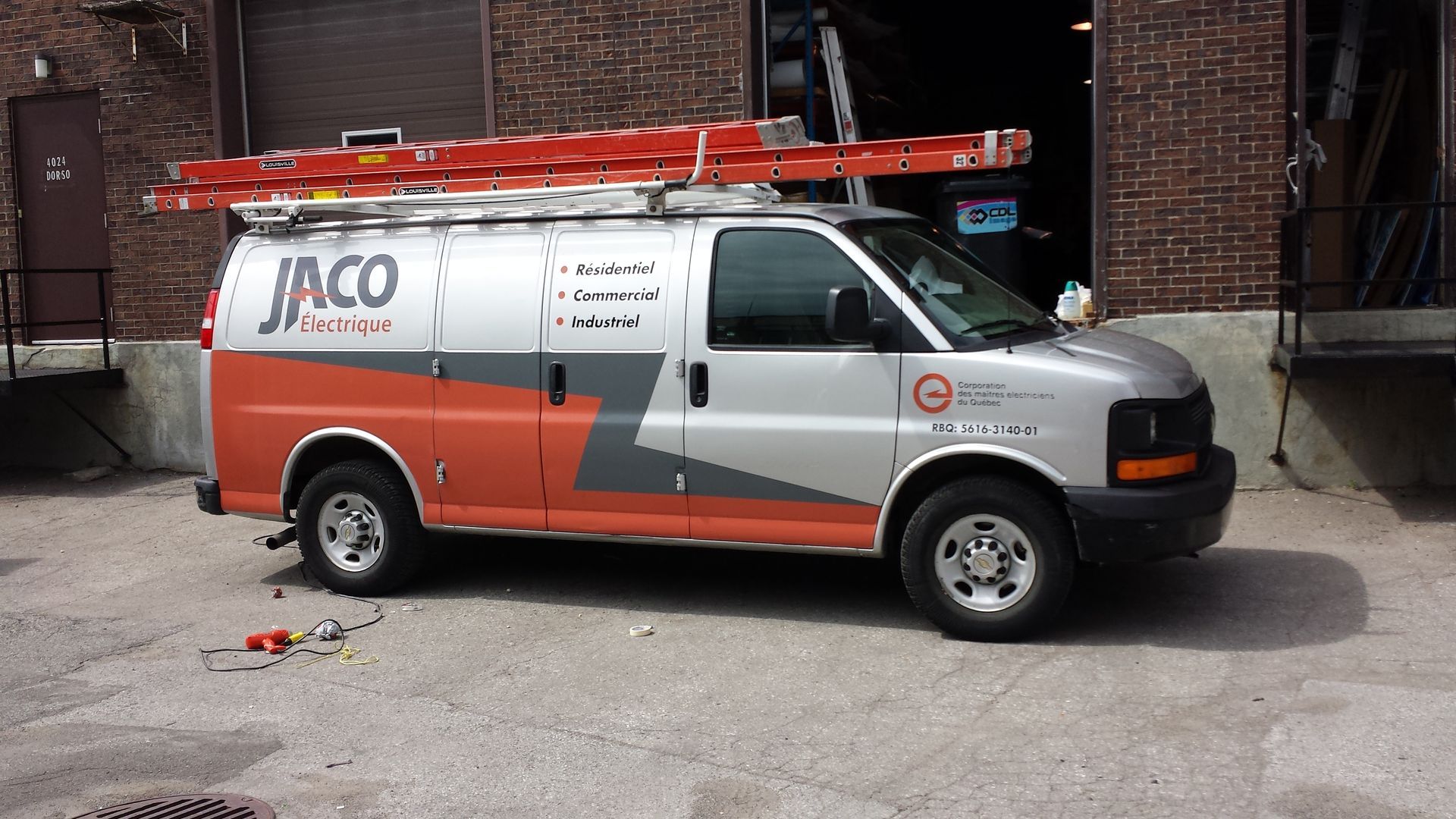 Silver van with orange accents, JACO Energy logo, parked near a brick building. Ladder on roof.