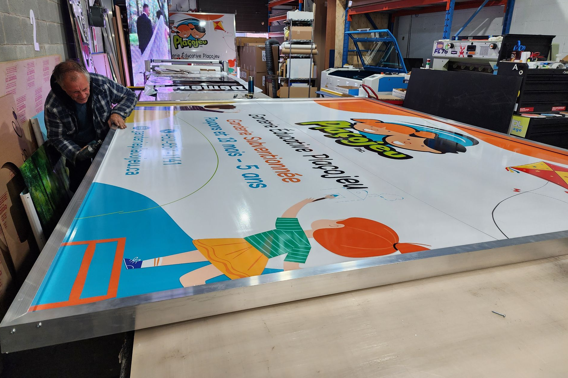 Man working on large colorful sign. Factory setting, orange, blue, and green graphics.
